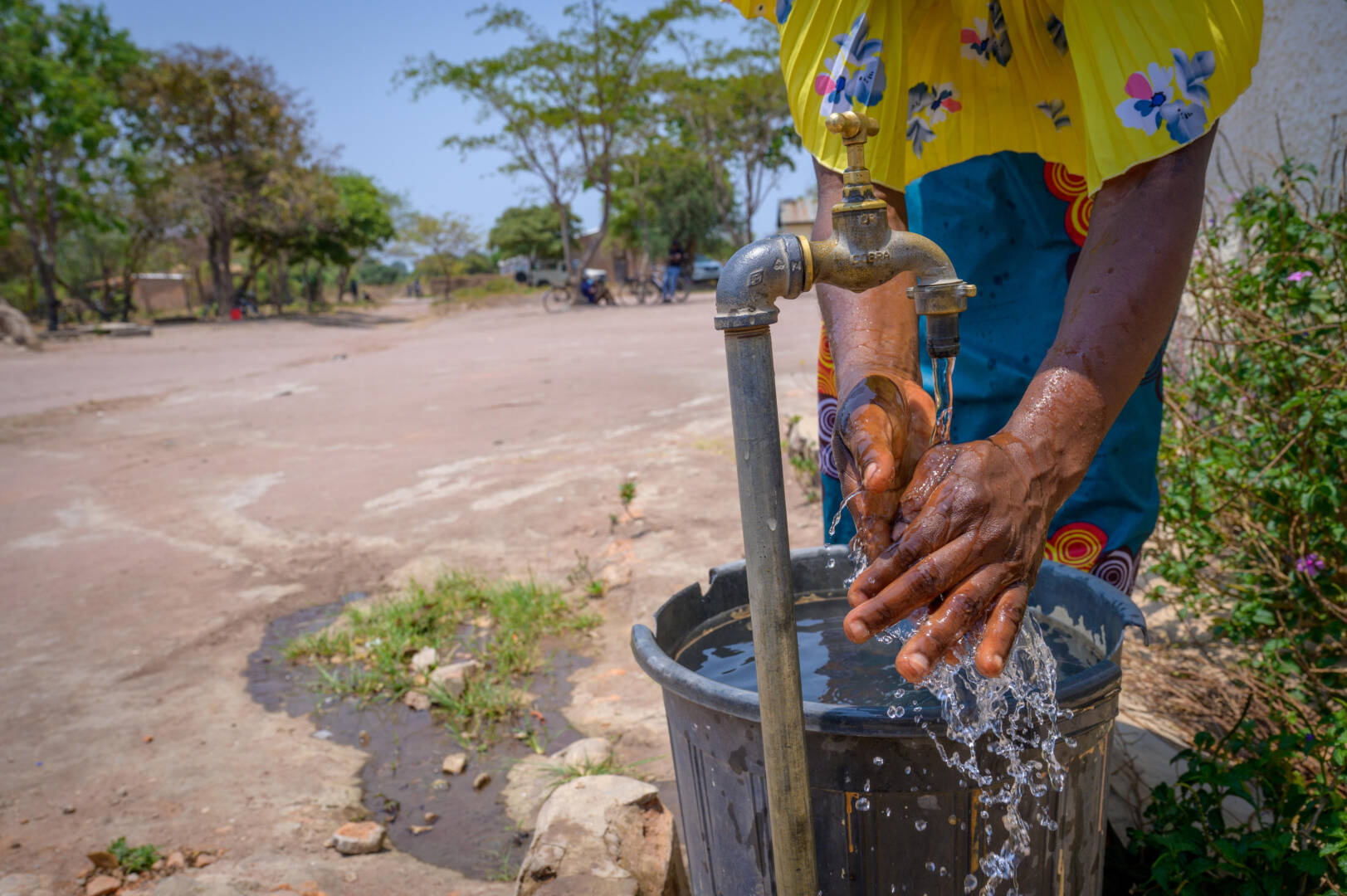 A woman rinsing her hands under a metal water tap. Water flows, splashing over a full bucket under the tap.