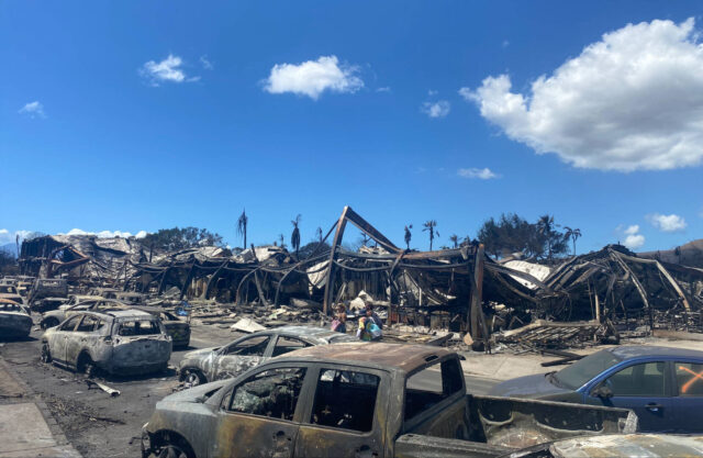 Cars, buildings, and homes are completely charred in contrast to a blue sky with several clouds.