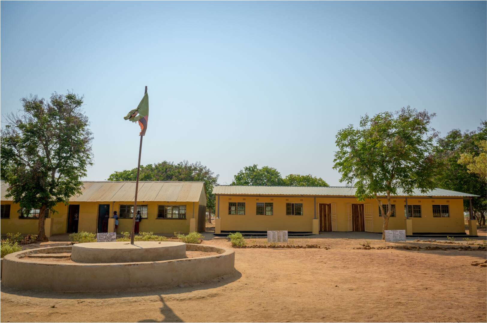 A Zambian flag waves on a pole in front of two single-story, tin-roofed school buildings. The sky behind the school is blue and clear.