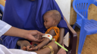 A young child sits on a woman’s lap while the circumference of their arm is measured. The tape shows a red color.