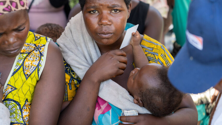 A mother gazes into the camera as her young son, wrapped in a baby sling, plays with a white card, holding it to her face.