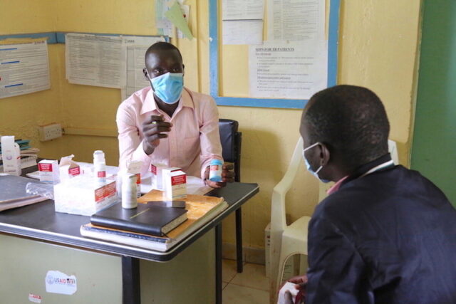 A man sits at a desk in a health clinic. He holds a bottle of pills while talking to another man.