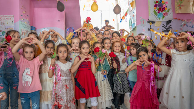 A large group of girls in a classroom shape their hands into hearts, while a few girls hold roses.