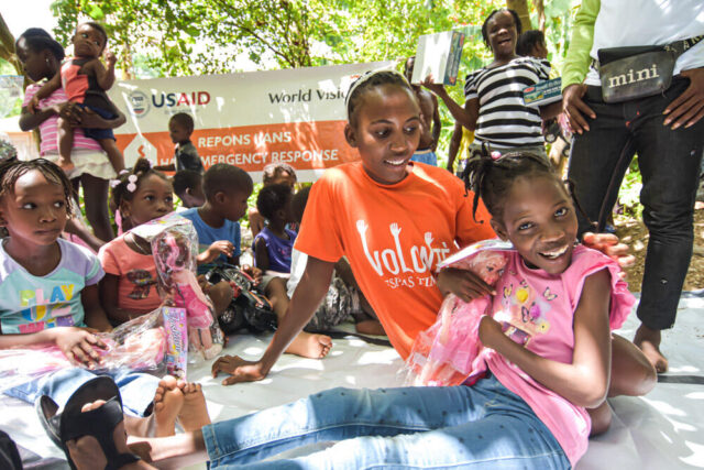 A child in a pink shirt smiles joyfully while hugging a doll. Kids and adults surround her. A USAID and World Vision banner is visible among trees.