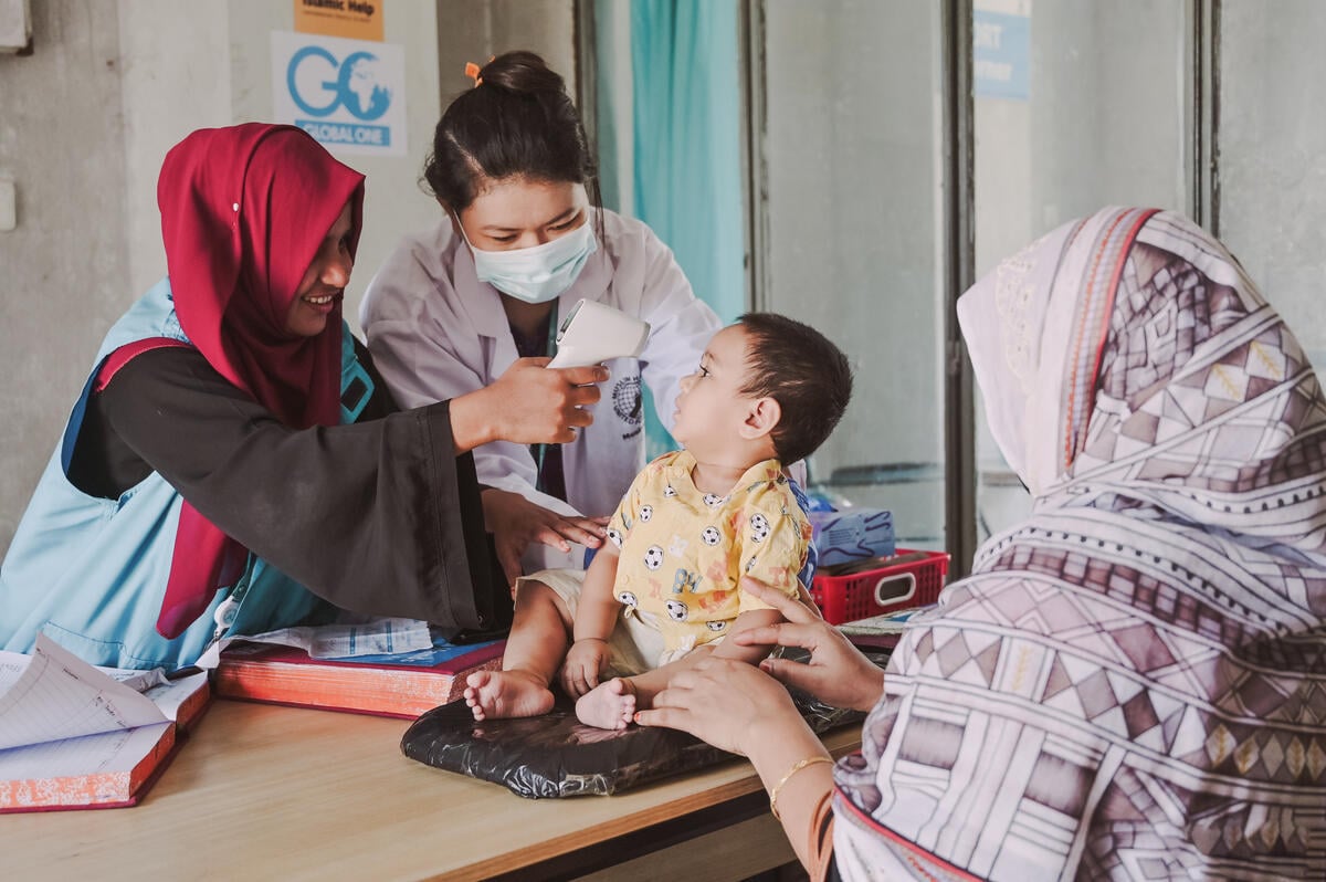 Health workers examine a young child seated on a mat in a clinic. A woman in a headscarf gently steadies the child.