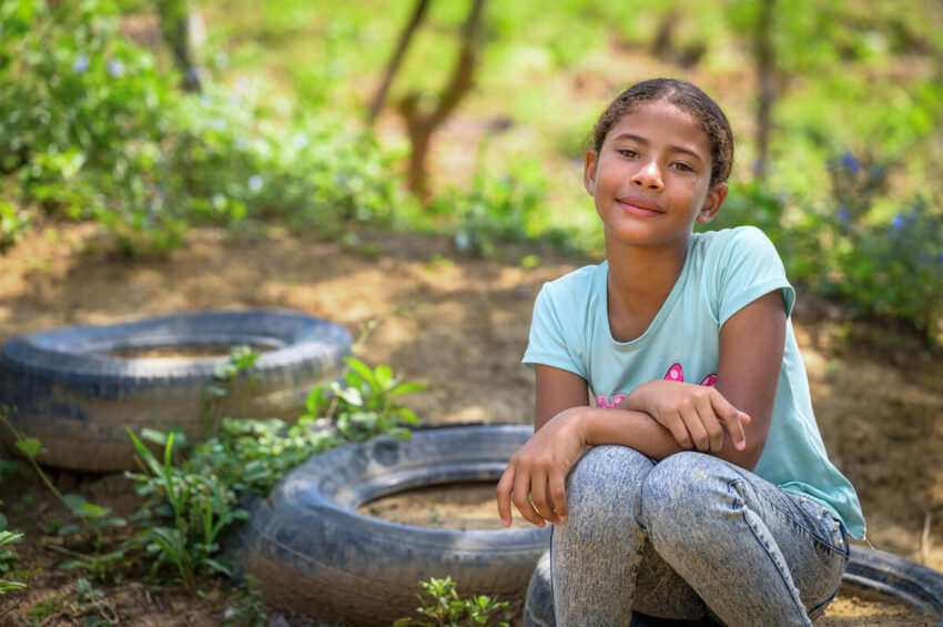 A young girl in a T-shirt and faded jeans sits on a tire on the ground and gazes into the camera.
