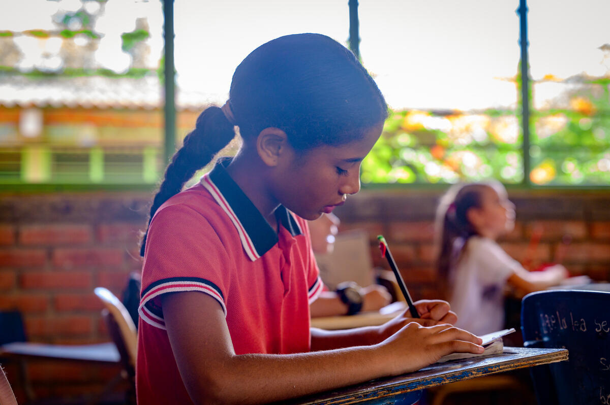 A focused young girl with a ponytail braid and red polo shirt concentrates on her schoolwork, holding a pencil at her desk in a classroom. 