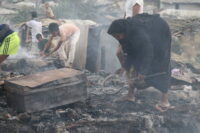 A woman searches through fire-ravaged debris. In the background, other people sift through the debris amid dust and smoke.