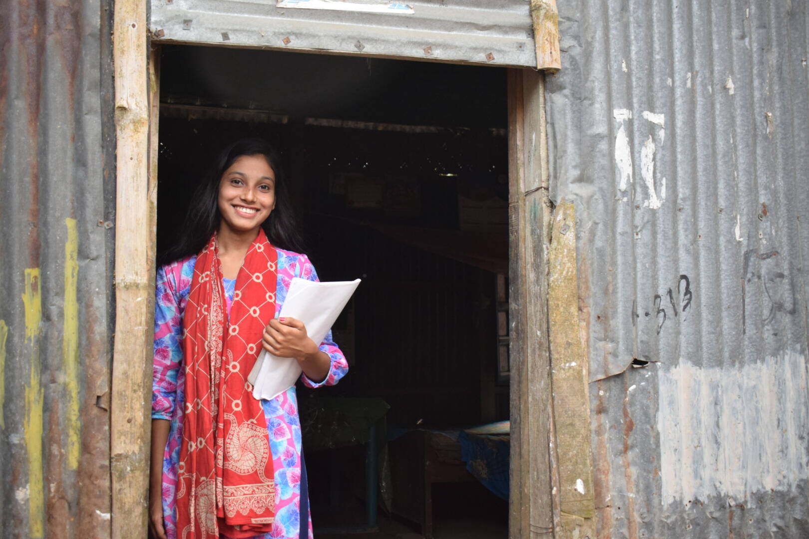 A teenage girl smiles widely in brightly patterned clothes and holds white papers in her hand.
