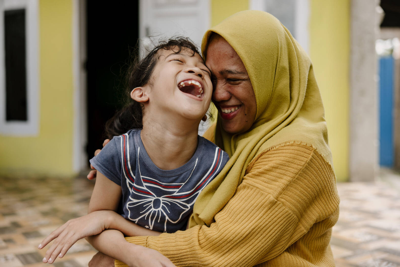 A smiling woman in a headscarf hugs a laughing girl.