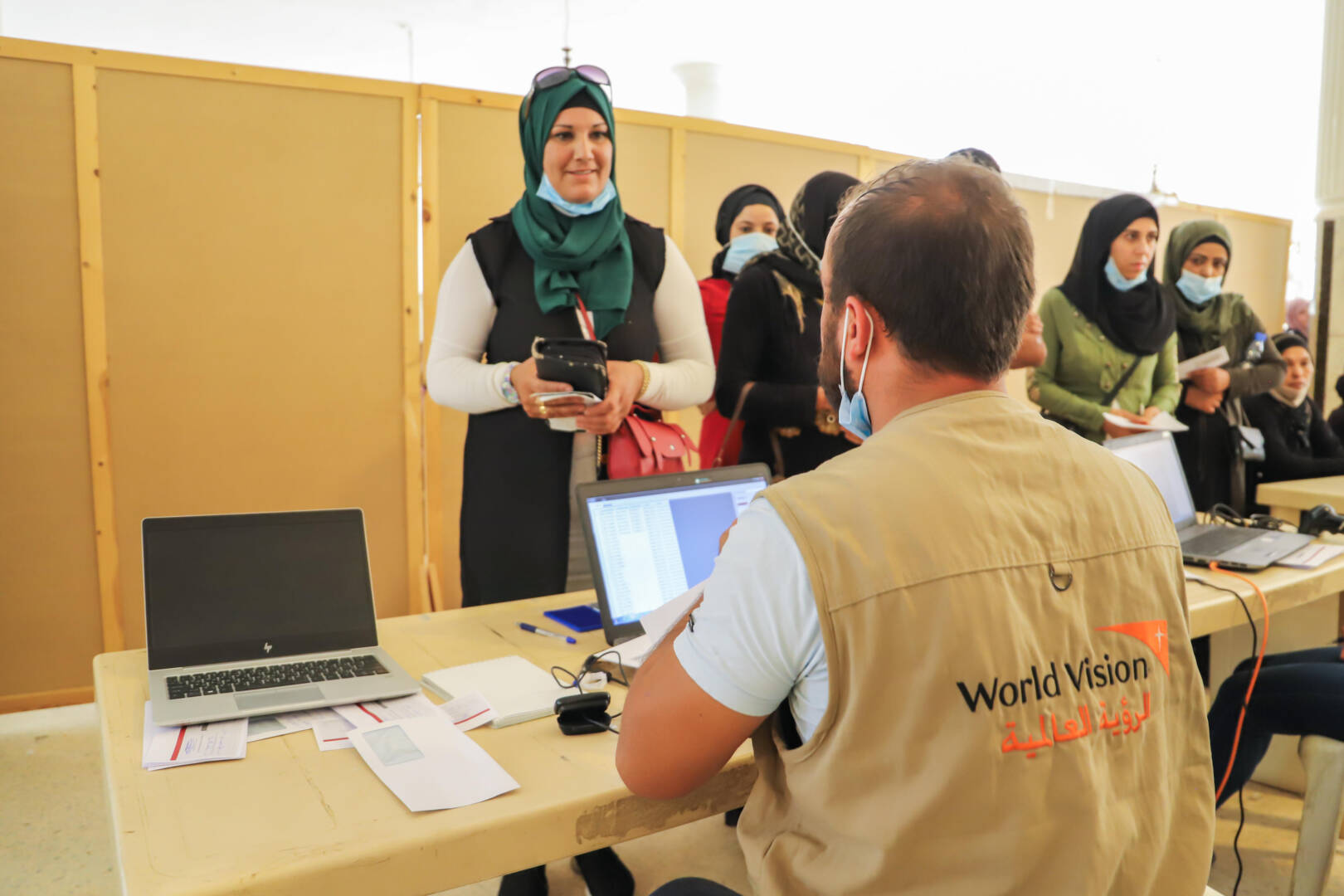 A man wearing a World Vision vest takes notes on a computer while a woman standing across the table from him speaks.