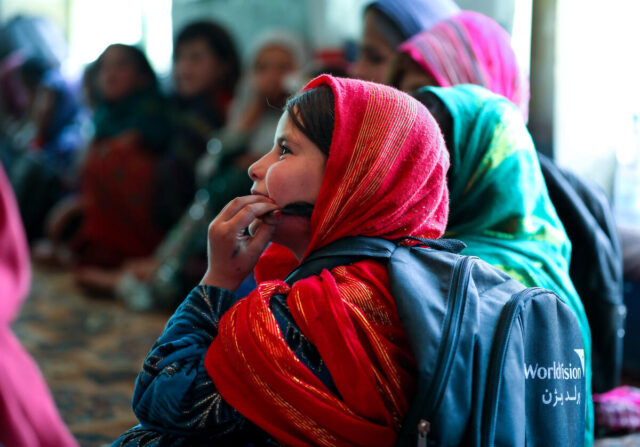 A child in a red headscarf gazes thoughtfully upward, holding a strand of hair close to her cheek. Kids are in the background.