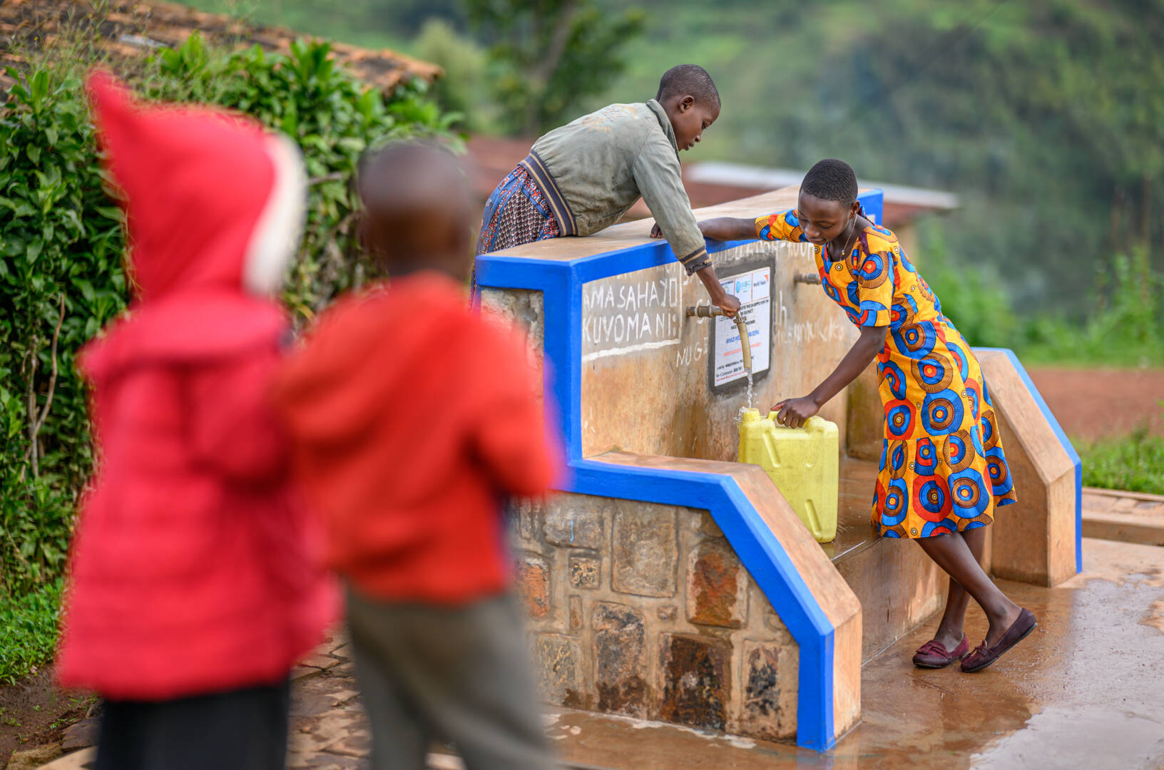 A Rwandan girl in a bright orange and blue dress fills her yellow jerrycan with water at a tap. She is helped by another girl leaning over the top.