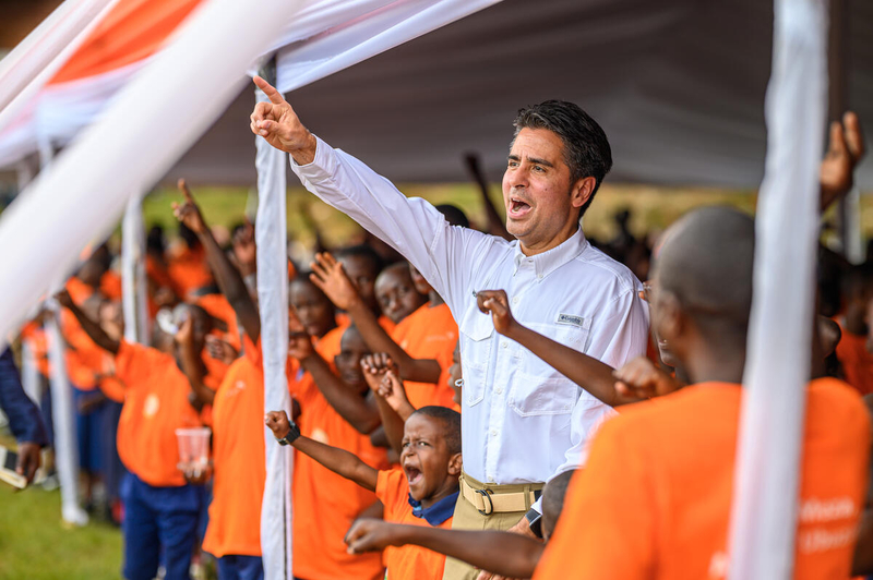 A man wearing white is surrounded by children wearing orange as they cheer with hands in the air.