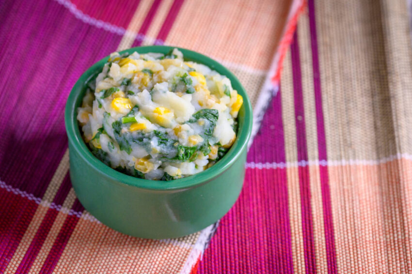 Mukimo, a mashed potato dish made with corn, peas, and spinach, sits in green ceramic dish.