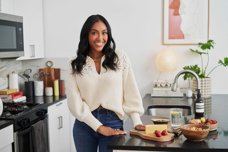 A woman wearing jeans and a cream sweater stands in a kitchen next to a wood board with cheese and strawberries on it.