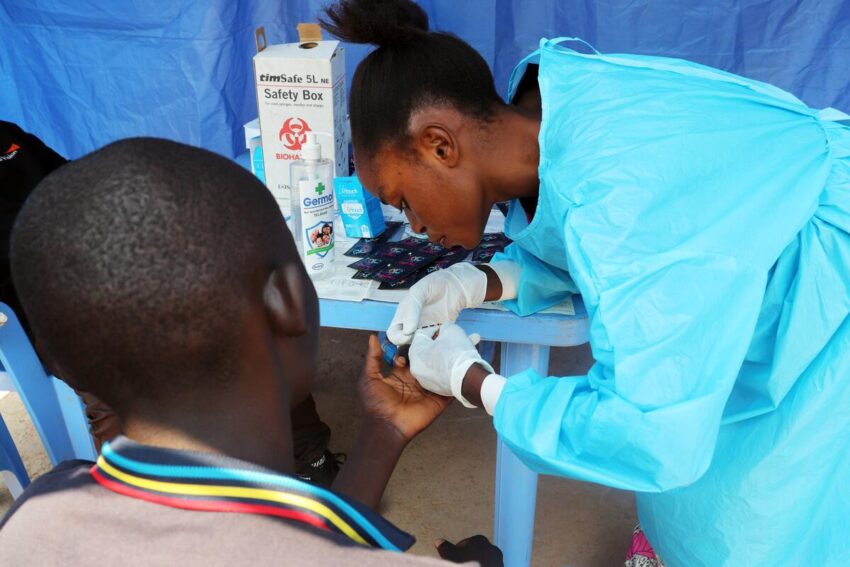 A woman in a blue medical gown and white gloves leans over a table with healthcare supplies, preparing a man’s hand during a screening in a mobile center.