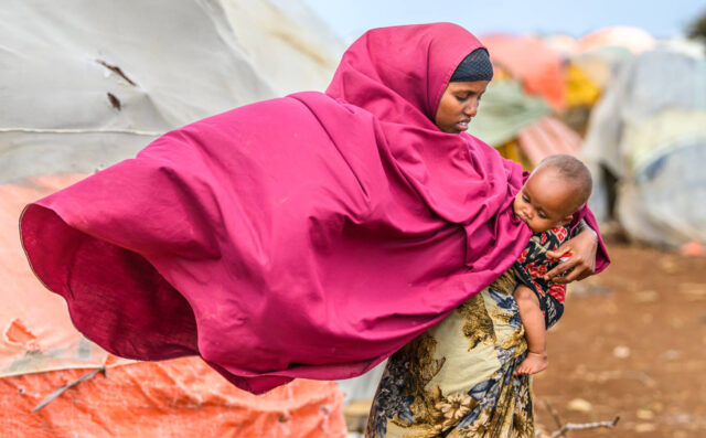 A woman in Somalia holds her baby amid canvas tents, her pink hijab billowing from a gust of wind.