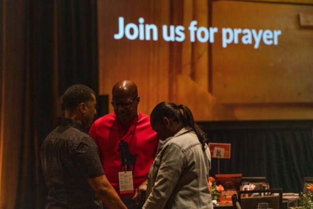 Three people stand in a circle, holding hands and praying. On the wall behind them the words “Join us for prayer” are projected.