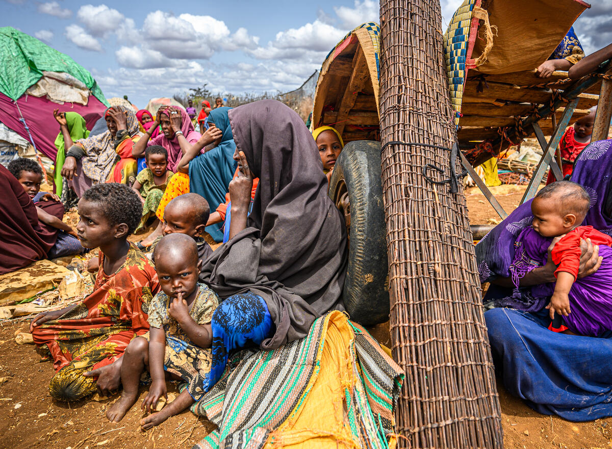 A veiled woman sits against a tire, her face turned in profile. Women and children surround her in a crowded camp.