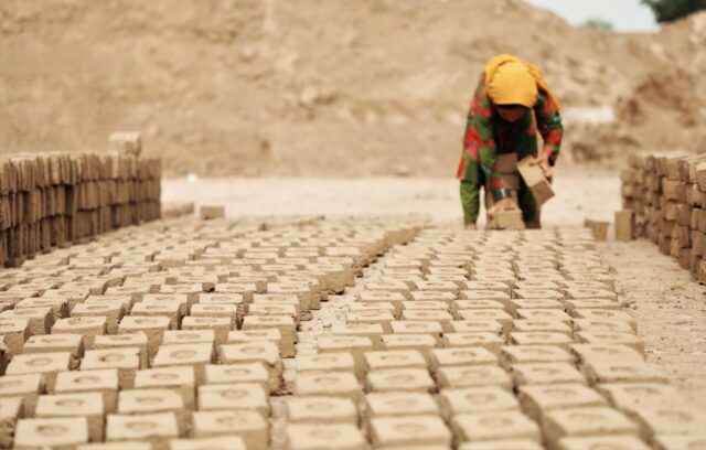 A young girl in a yellow headscarf and red-green patterned outfit arranges bricks in several rows outside a factory in Afghanistan.