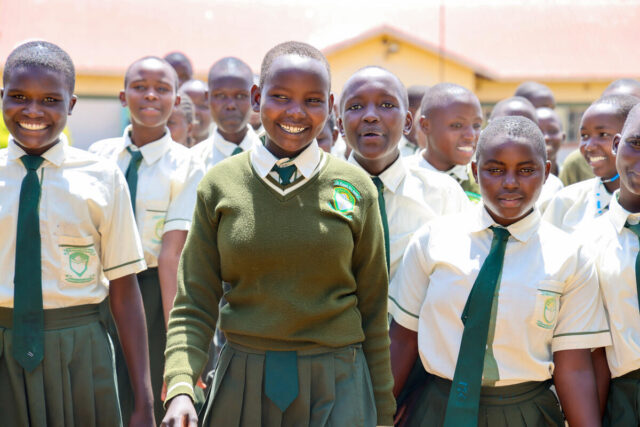 Smiling schoolgirls dressed in white shirts, green ties, and matching skirts walk in a group from a building with a pink roof, bathed in sunshine.