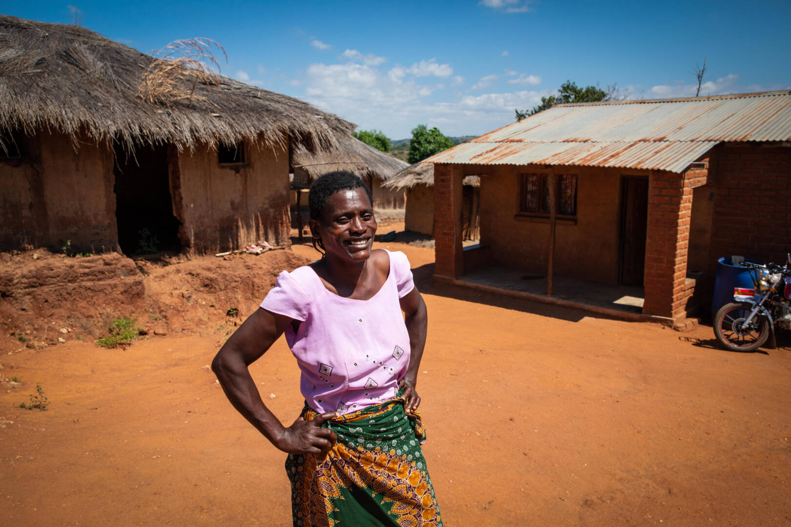 A woman stands, hands on her hips. Behind her is a mud hut with a thatched roof, and a newer building made of brick.