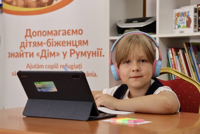 A young blonde girl faces the camera, seated in a learning space. She’s wearing headphones, and an open laptop sits before her.