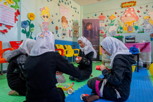 Four girls in white veils sit on the floor in a circle, working on crafts in a brightly colorful room.