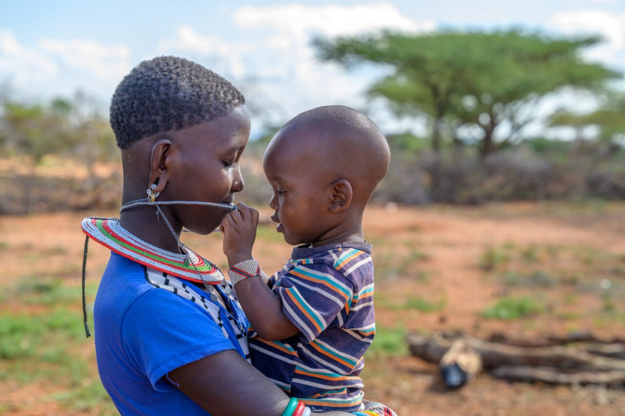 A young woman holds a young boy in her arms. The child plays with the young woman’s beaded necklace.
