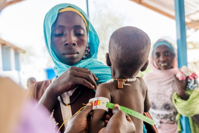 A woman holds her young child while a health worker measures the child’s arm for malnutrition. The measuring tape shows red.