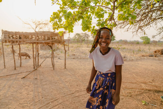 A joyful girl smiles widely in a blue patterned skirt in a dry, rural landscape.