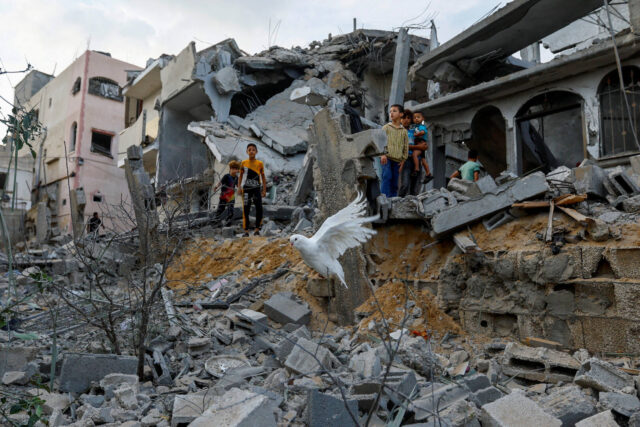 A dove flies in the foreground as children stand among the debris of houses destroyed in a conflict.