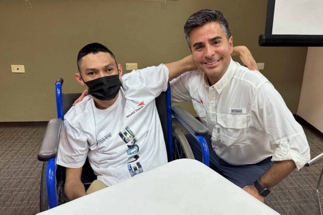 A young man in a white T-shirt sits in a wheelchair with his arm around a middle-aged man in a casual white button-down shirt.