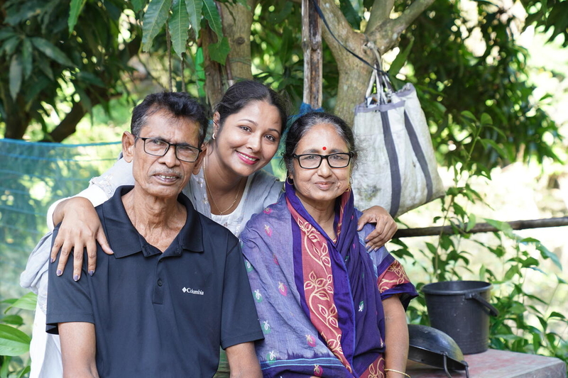A young woman stands with her arms around two older people who are sitting. They are all smiling. There are trees behind them.
