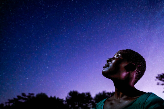 A young woman looks up at an indigo-colored night sky full of stars.