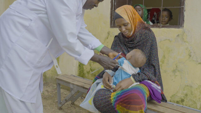 A health worker holds paper tape and a pen while reaching for a baby in a mother’s lap.