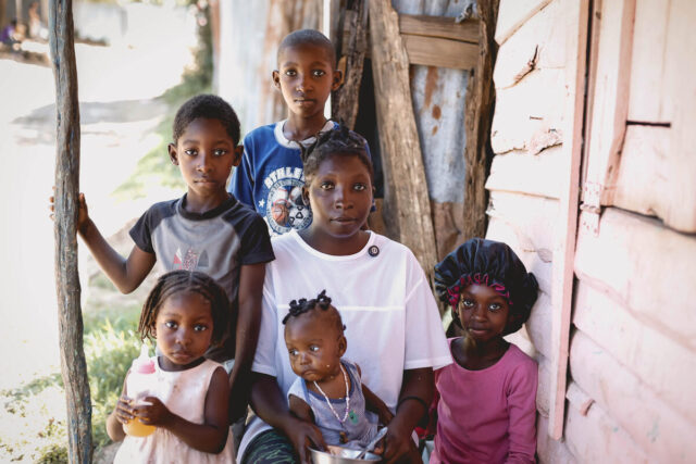 A woman in a white shirt gazes into the camera, surrounded by five young children. Four of the children look directly at the camera as they sit outside by a wooden structure.