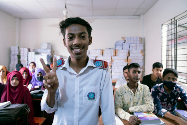 A smiling male student in a white shirt gives a peace sign in a classroom with peers in the background.
