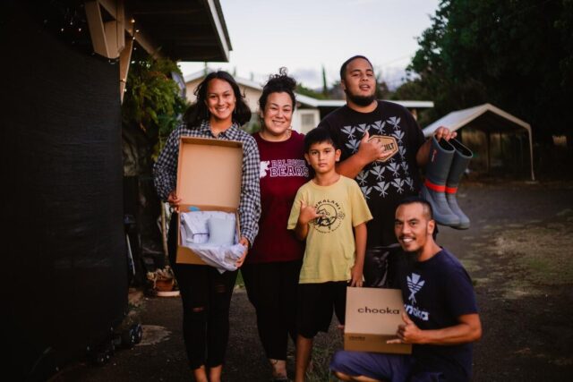 A family of five smiles at the camera. Three family members hold donated boots, and three males make the Hawaiian shaka sign with their hands.