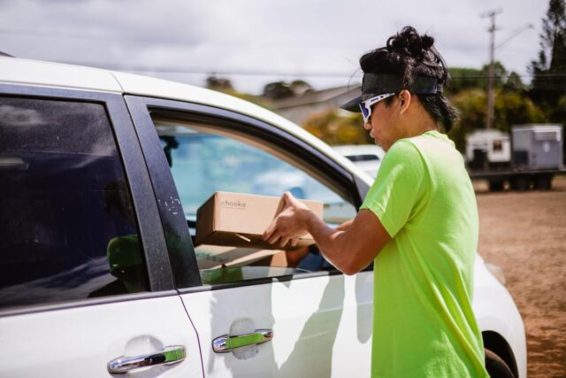 A volunteer in bright green T-shirt and white sunglasses hands a box of shoes to a passenger in a white vehicle.