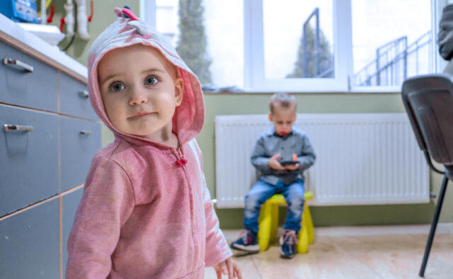 A young girl with blue eyes in a pink hooded sweatshirt glances sideways as a boy appears in the background.