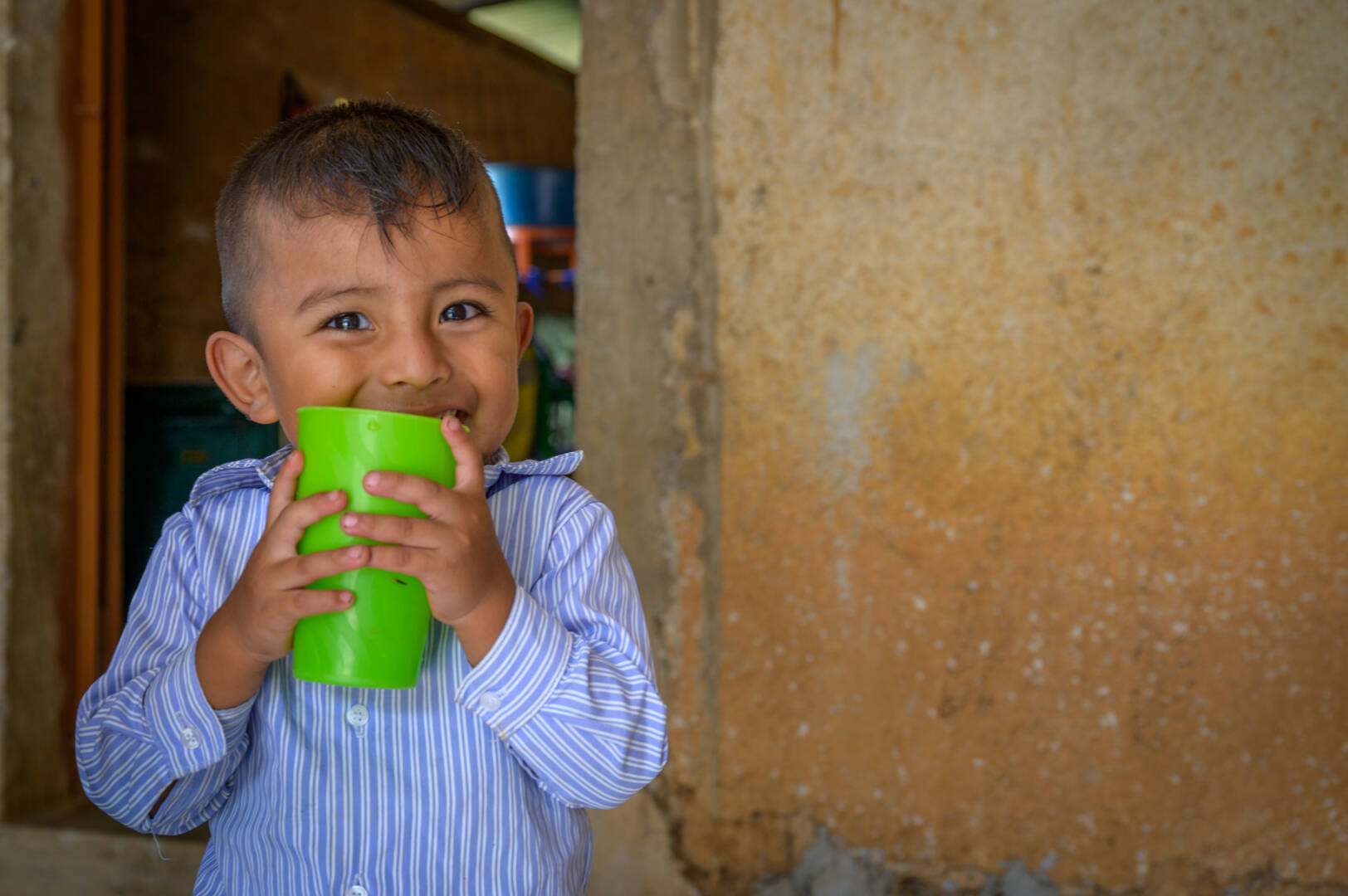 A boy smiles at the camera as he holds a green cup up to his lips.