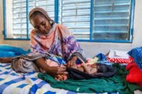 A woman looks down lovingly and gently holds her baby’s legs as the child lies on a blanket on a bed beneath a window with blinds.