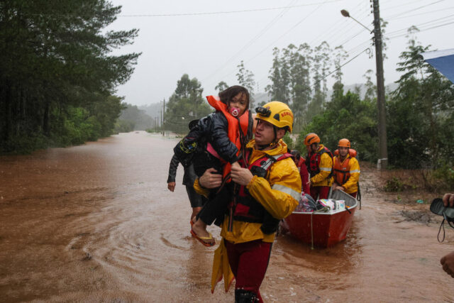 A man in a yellow hard hat carries a young girl with a pacifier and life vest through floodwaters, with a rescue team and boat behind them.