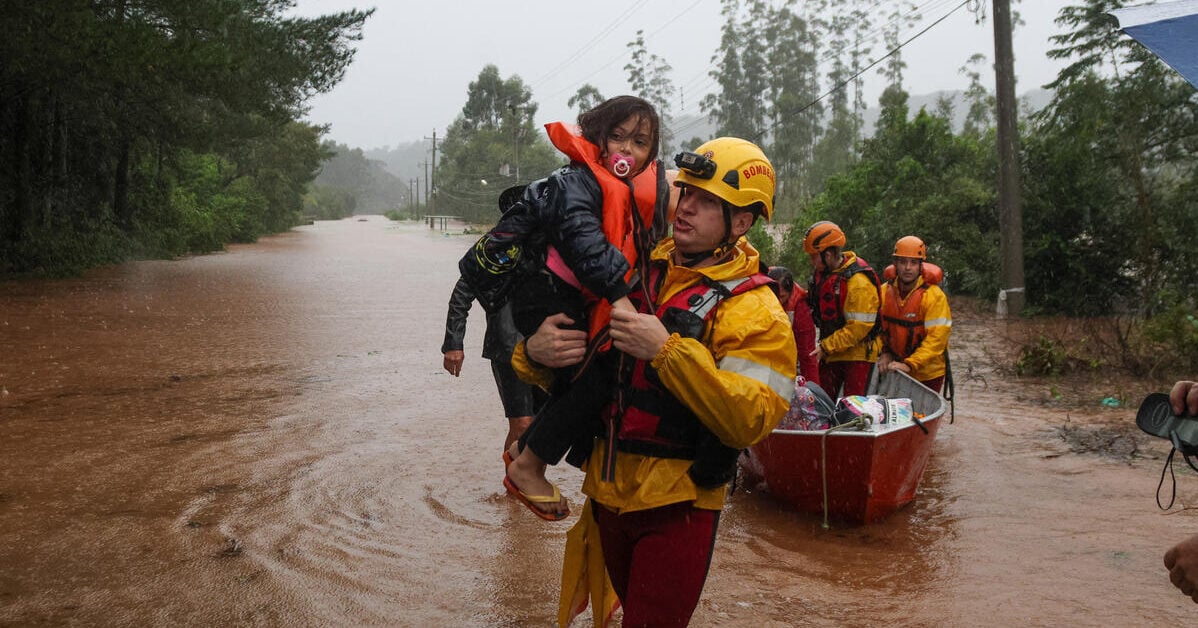 A man in a yellow hard hat carries a young girl with a pacifier and life vest through floodwaters, with a rescue team and boat behind them.