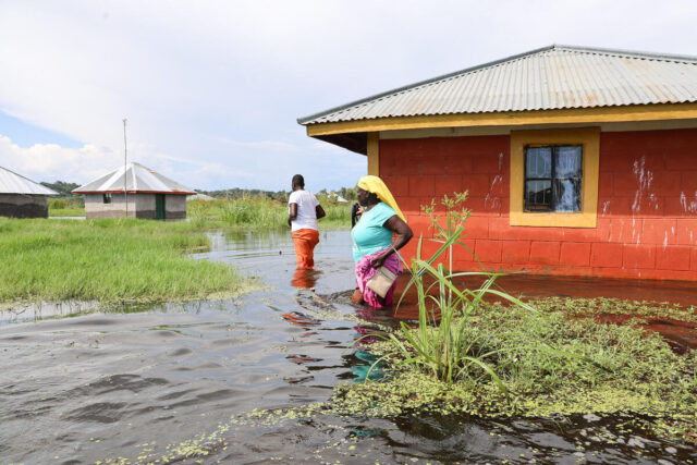 A man and a woman wade through floodwaters next to a house painted orange and yellow, with other structures visible in the distance.