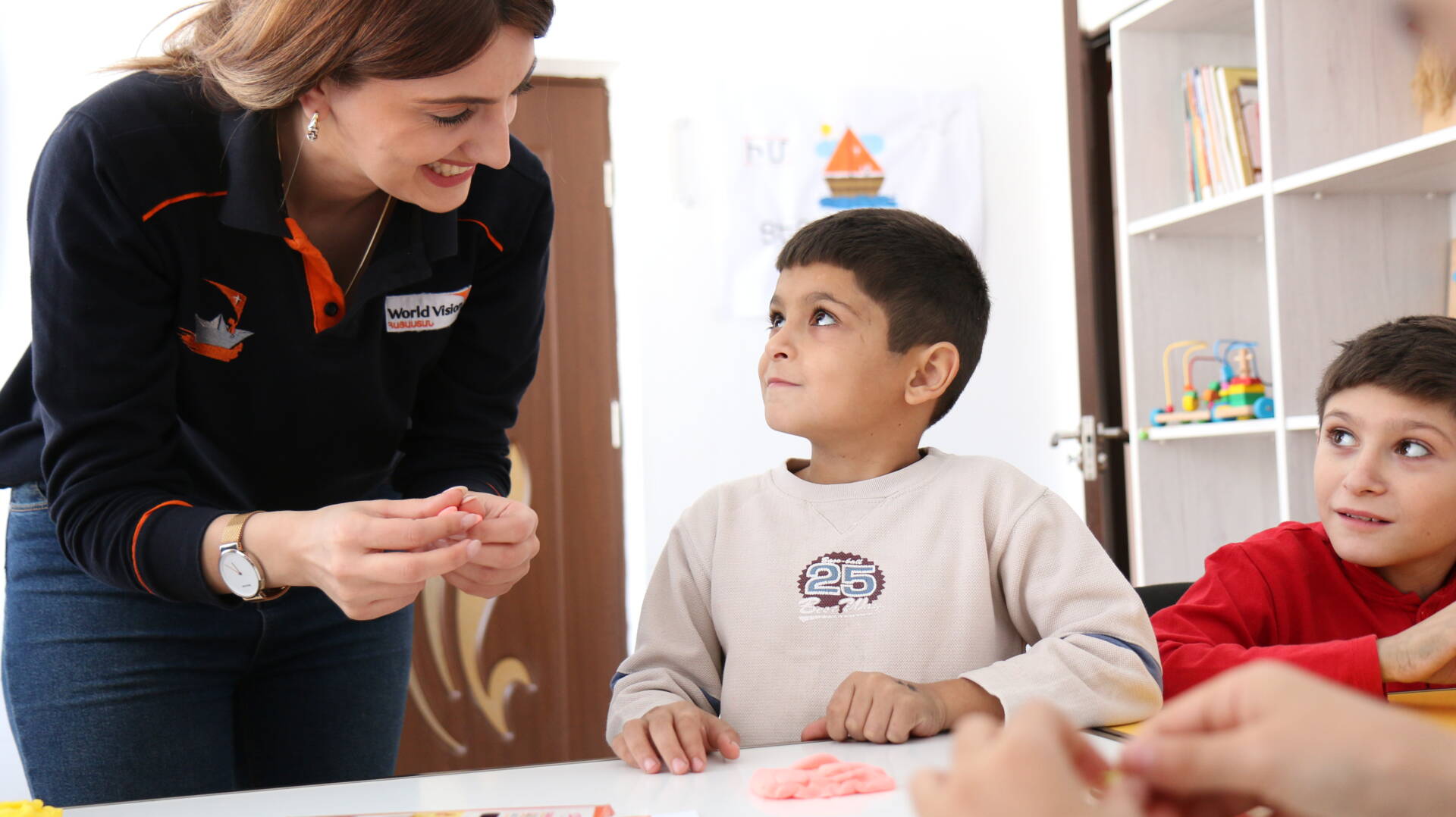 A woman smiles as she leans over a table where two boys sit looking up at her. She holds a piece of playdough in her hands.