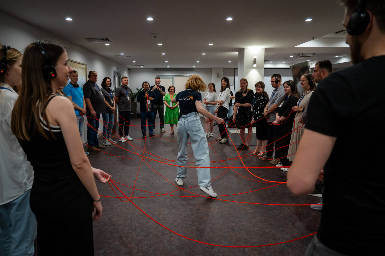 A group of people stand in a large circle, each holding on to a long strand of red yarn forming a web across the circle. One woman, wearing a World Vision shirt, stands in the center holding the yarn.