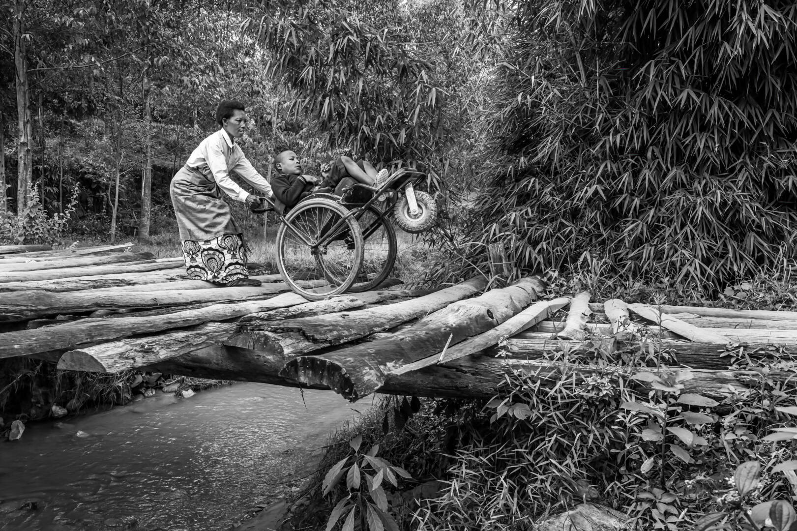 Black-and-white photo of an African woman pushing a boy in a wheelchair across a wooden bridge.